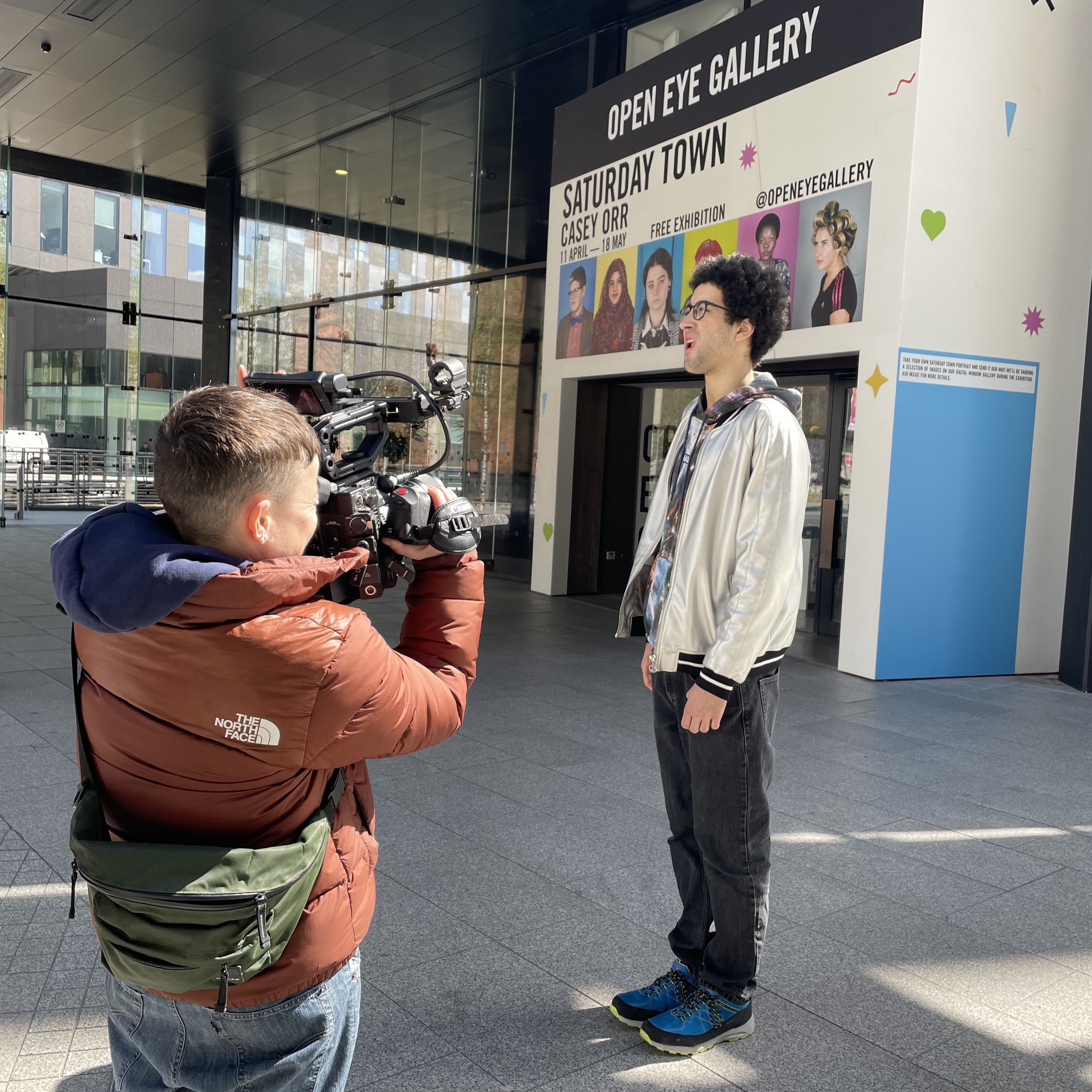 Patrick Bannon, a RAWD associate artist, stood outside the Open Eye gallery. He wears a silver jacket, with a NASA hoodie, He is being filmed by Jay