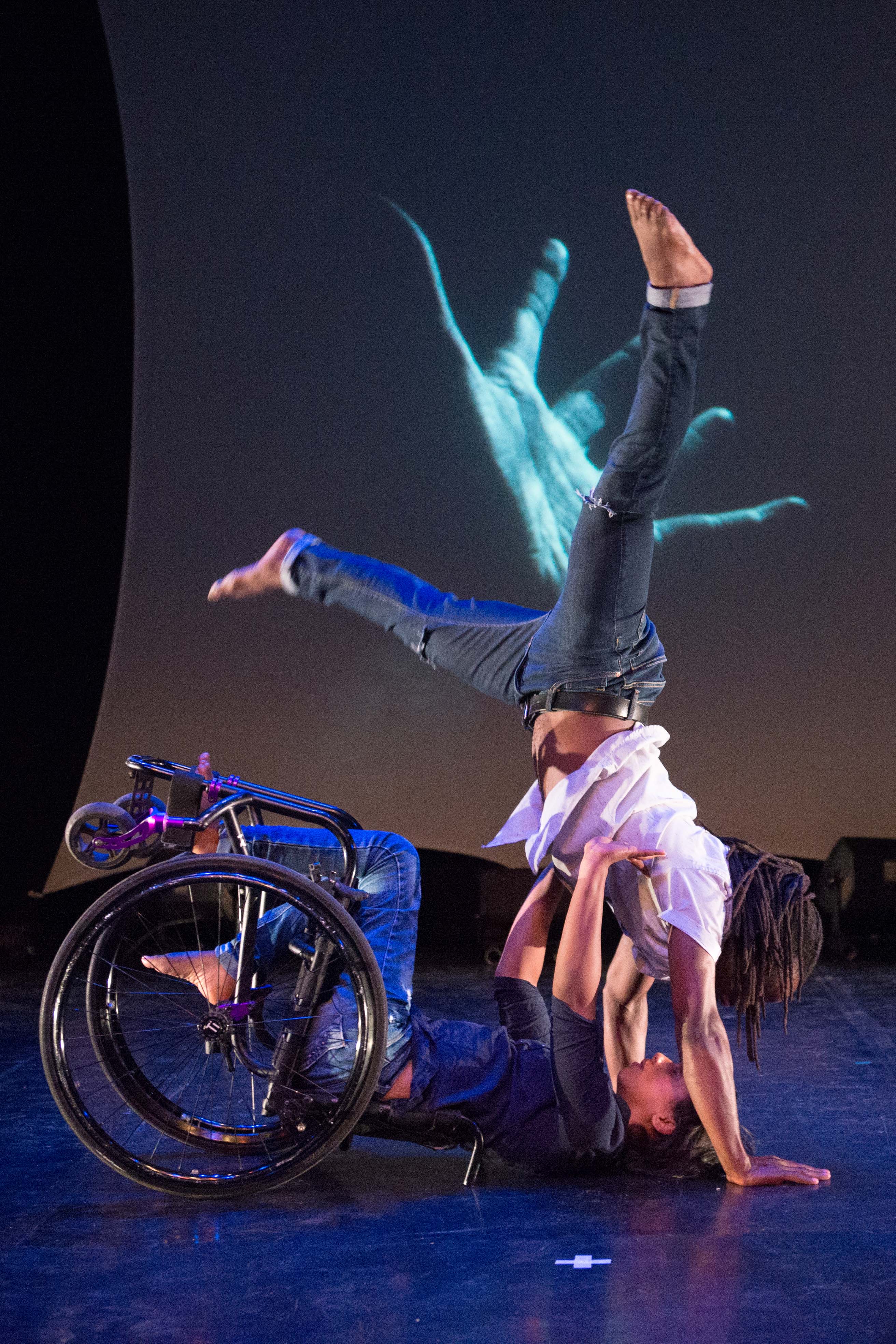 Dancer in jeans and white t-shirt carries dancer in wheelchair held sideways towards a screen showing a large image of a hand in teal