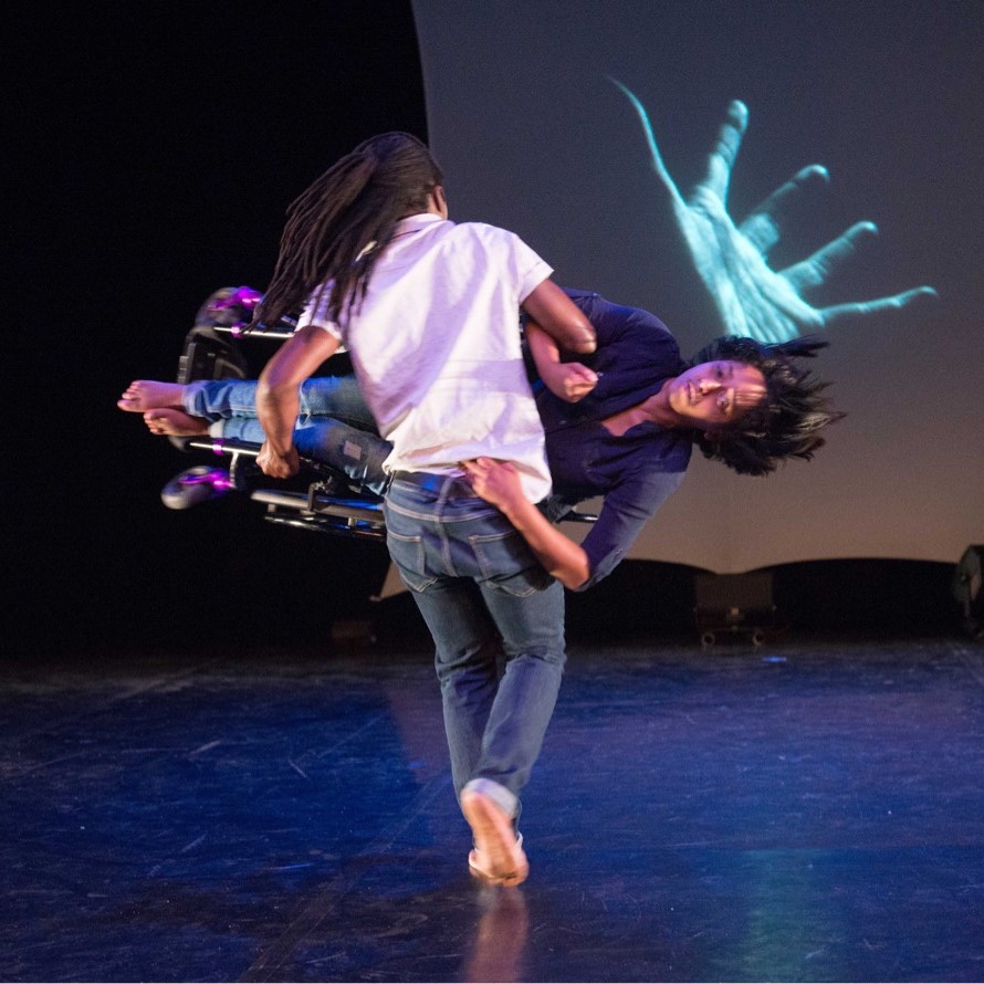 Dancer in jeans and white t-shirt carries dancer in wheelchair held sideways towards a screen showing a large image of a hand in teal
