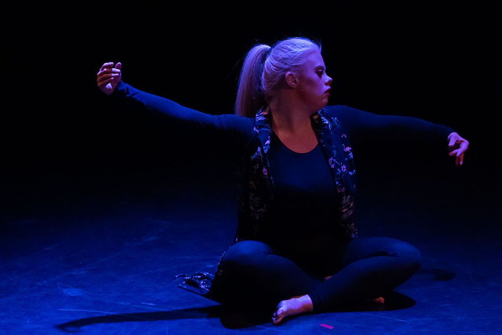 Helen Cherry, a fair-haired dancer in a black leotard, sits cross-legged onstage, spreading her arms gracefully. 