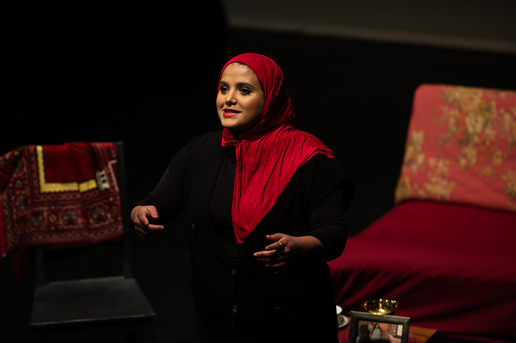 Amina Atiq wearing a black outfit and red hijab addressing an audience earnestly. 