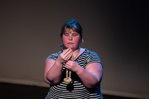 Bethany Murray, a performer wearing a black and white striped top, stands onstage, transfixed by a small wooden mannequin she holds before her. 