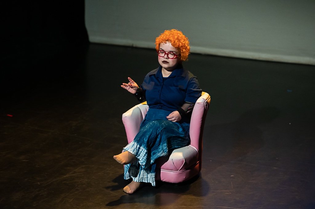 Tammy Reynolds, a performer with short, orange, curly hair, sits on a small pink arm-chair. She has her eyes closed and appears to be listening to something. 