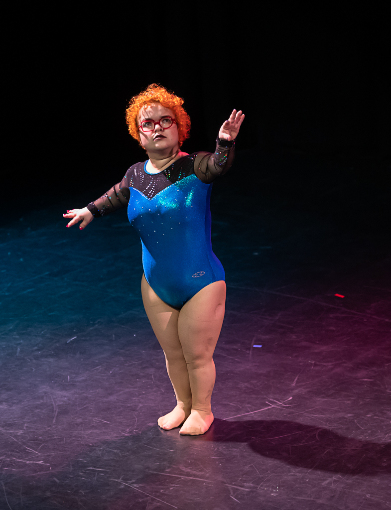 Tammy Reynolds, a performer with short, orange, curly hair, raises one arm gracefully whilst dressed in a shiny blue leotard. 