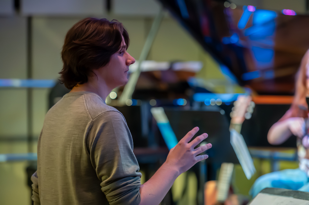 Thomas, a teenage boy with flowing dark hair just above the shoulder wear a green jumper. He is facing away from the camera and conducting an orchestra who are out of shot