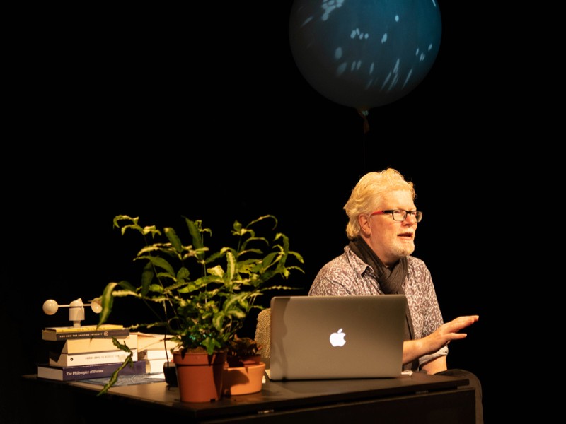 Aidan Moesby sitting at a desk, apparently on stage as he addresses someone unseen. A balloon is above him.