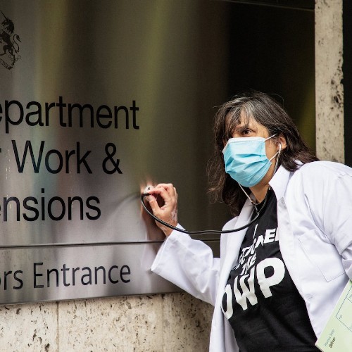 Dolly sens in doctor coat and mask putting a thermometer against the sign of the Department of Work and Pensions