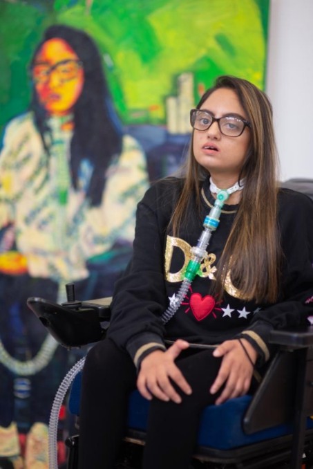 Artist Juvairiyya Patel sits in front of a portrait painting of herself. She wears glasses and has long hair.