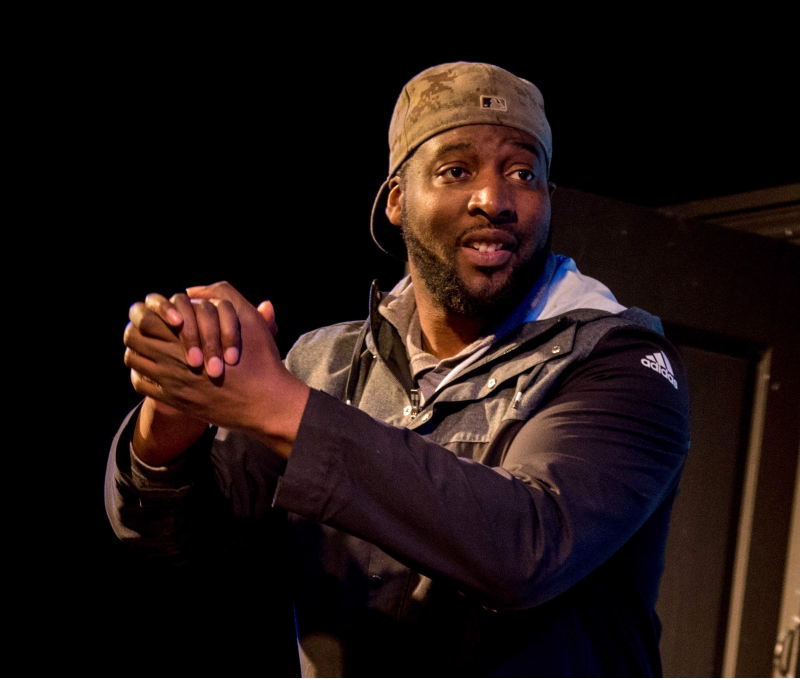 A man clasping his hands together, wearing a hat in a dark space