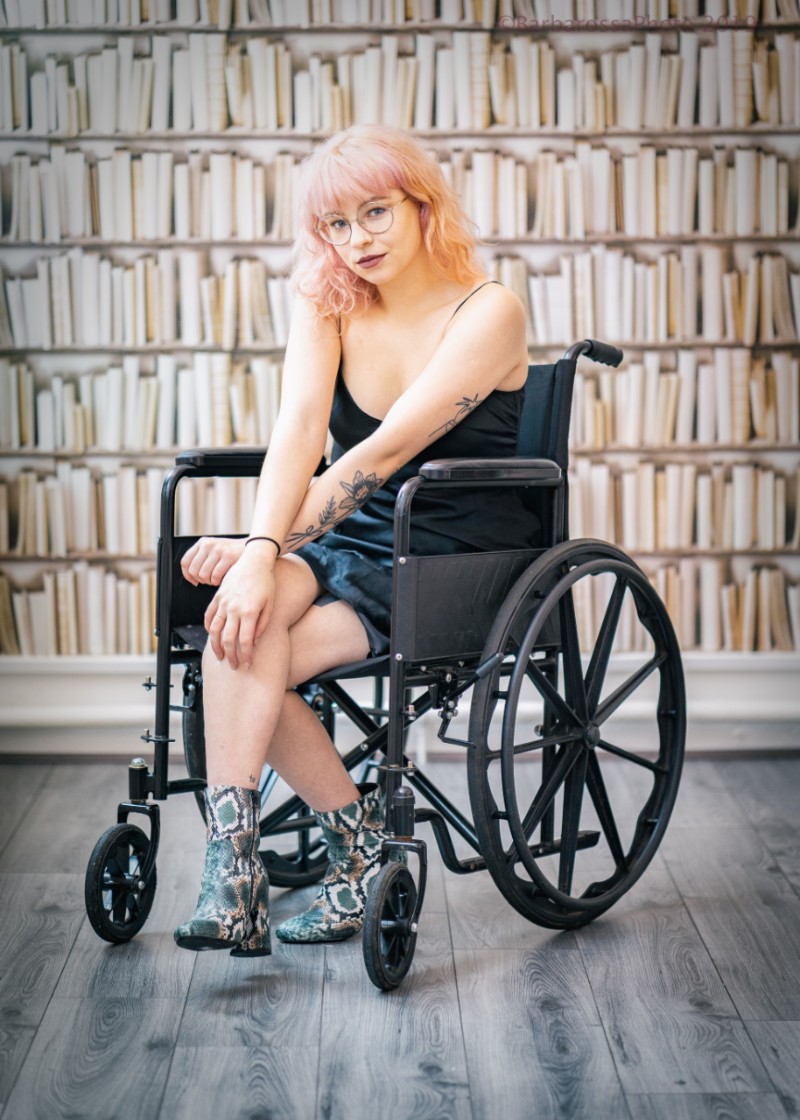 Photographer Natalie Bedkowska photographed sitting in her wheelchair in front of bookshelves. She wear glasses and has pink hair.