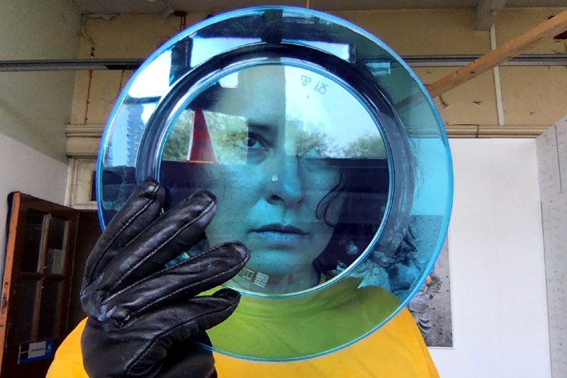 Nicola looks through a transparent blue plate holding the plate with a black leather glove, she looks dreamily off screen. 