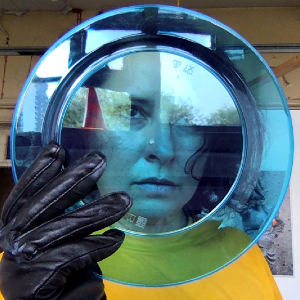 Nicola looks through a transparent blue plate holding the plate with a black leather glove, she looks dreamily off screen. 