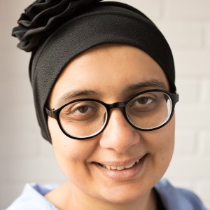 A smiling, British Asian woman wearing glasses and a blue shirt. 