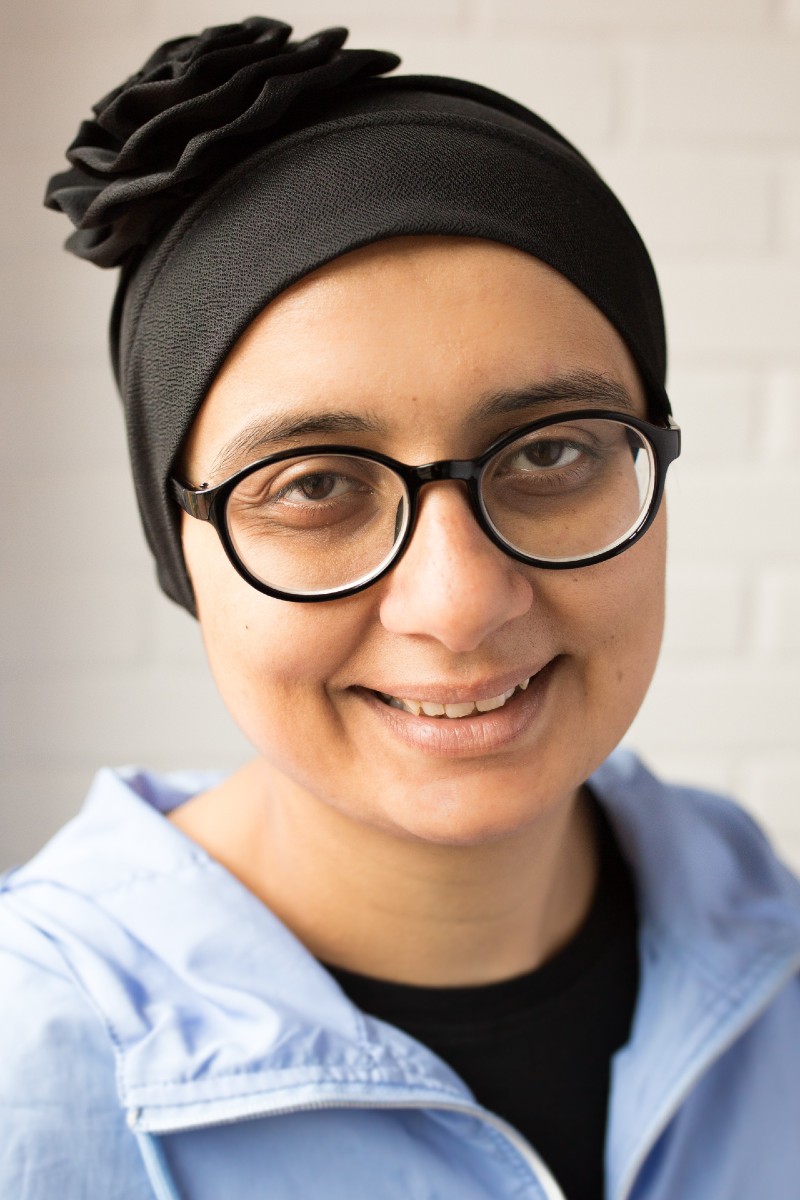 A smiling, British Asian woman wearing glasses and a blue shirt. 