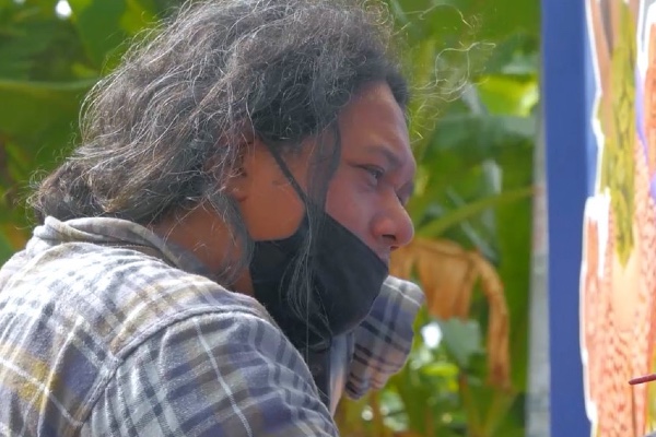 An Indonesian man in a greyish flannel shirt pictured in profile looking pensively at something. He wears a black PPE mask