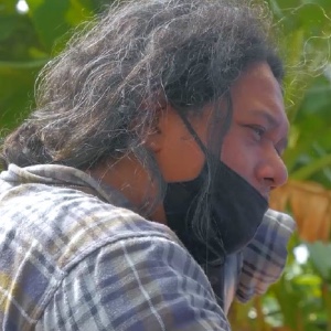 An Indonesian man in a greyish flannel shirt pictured in profile looking pensively at something. He wears a black PPE mask.