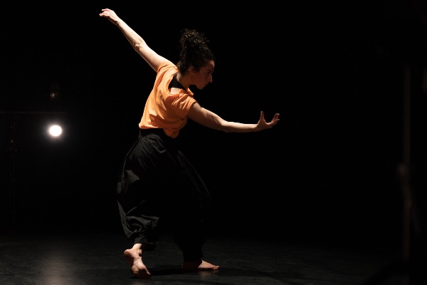 A dancer in a dark performance space wearing an orange top and black trousers, pictured side-on with arms outstretched