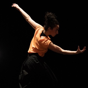 A dancer in a dark performance space wearing an orange top and black trousers, pictured side-on with arms outstretched