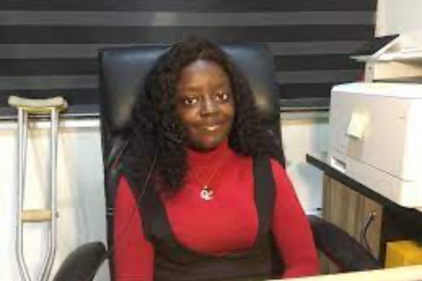 A young Nigerian woman with long black hair sat at a desk smiling wearing a necklace and a red top  