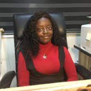 A young Nigerian woman with long black hair sat at a desk smiling wearing a necklace and a red top 