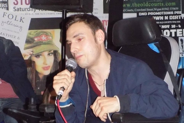 A Caucasian man speaking into a microphone. He has light brown hair, uses a wheelchair and wears a blue hoodie