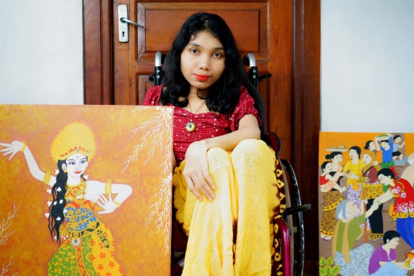 A Balinese woman with shoulder length black hair who is using a wheelchair and wears a red top and lipstick. Next to her are two colourful paintings