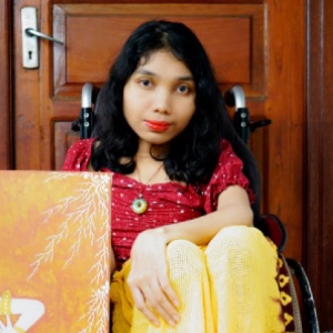 A Balinese woman with shoulder length black hair who is using a wheelchair and wears a red top and lipstick. Next to her are two colourful paintings
