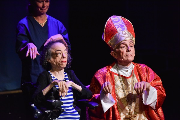 Woman in wheelchair on stage holding her hands in mid air next to a man dressed as a priest