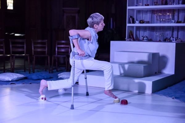 Woman on crutches striding across stage balancing feet on tea cups