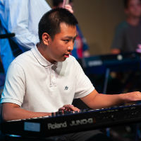 A photo of a young man dressed in a white short sleeved shirt on stage playing an electronic keyboard.