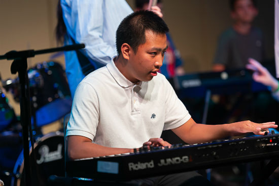 A photo of a young man dressed in a white short sleeved shirt on stage playing an electronic keyboard.