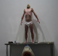 A man stands on a table wearing white underpants and a mask. Beneath the table is a plastic head of a shark.