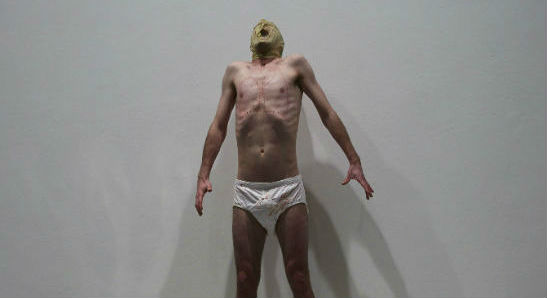 A man stands on a table wearing white underpants and a mask. Beneath the table is a plastic head of a shark.