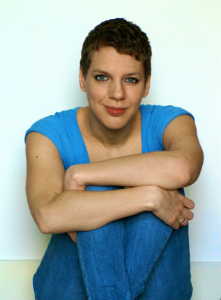 A photograph of Francesca Martinez sitting on the floor with her arms wrapped around her legs. She is dressed in blue jeans and a blue t-shirt.