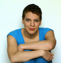 A photograph of Francesca Martinez sitting on the floor with her arms wrapped around her legs. She is dressed in blue jeans and a blue t-shirt.