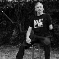 A black and white photo of Giles Duley sitting on a chair and wearing a t-shirt with the slogan Legacy of War Foundation