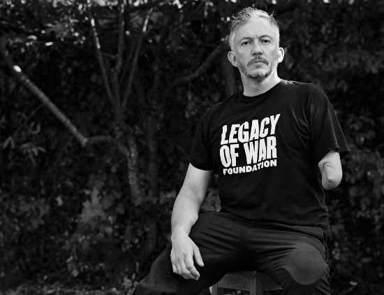 A black and white photo of Giles Duley sitting on a chair and wearing a t-shirt with the slogan Legacy of War Foundation