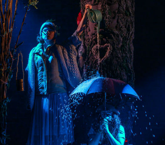 A photo of a man sitting on a dimly lit stage holding an umbrella over his head whilst a woman pours water on it from a watering can.