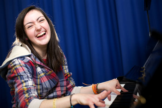 A young woman is playing a piano. She is turning towards the camera and smiling
