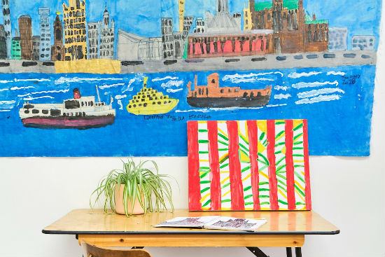 A photo of a plant and a painting sitting on a desk. Behind the desk on the white wall is a painting.