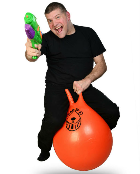 A man in a back t-shirt and trousers holds a green toy gun and bounces on an orange space hopper against a white bakground