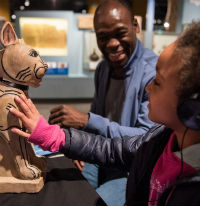 A photo of a man and a young girl sat at a table in front of a digital replica of an ancient Egyptian cat. The girl is wearing headphones. 