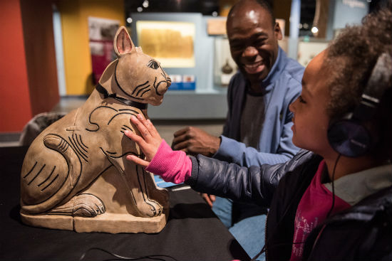 A photo of a man and a young girl sat at a table in front of a digital replica of an ancient Egyptian cat. The girl is wearing headphones. 