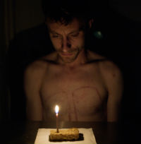 A photo of a bare chested man sitting in front of a table. On the table is a flapjack with one lit candle.