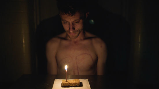 A photo of a bare chested man sitting in front of a table. On the table is a flapjack with one lit candle.