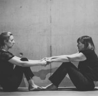 A black and white photo of two women sat on the floor across from each other. Their knees are bent, arms outstretched and they are holding hands.