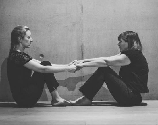 A black and white photo of two women sat on the floor across from each other. Their knees are bent, arms outstretched and they are holding hands.