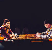 Two men wearing high visibility vests and woolly hats sit across a table from each other.