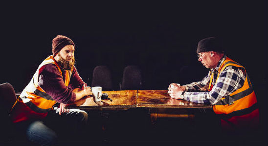 Two men wearing high visibility vests and woolly hats sit across a table from each other.