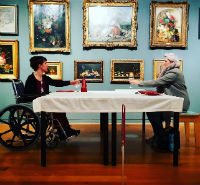 A woman sits across a table from a man in a wheelchair. They are raising a glass to each other. Behind them is a wall covered in paintings.